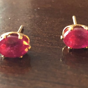 May include: A pair of gold-toned stud earrings, each featuring a vibrant, oval-shaped, red gemstone. The gemstones are set in a decorative, scalloped bezel. The earrings are displayed on a dark brown surface.