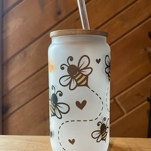 May include: A frosted glass tumbler with a bamboo lid and a clear drinking straw. The tumbler has a design of bees and hearts on a white background.