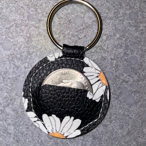May include: A black leather keychain with a white daisy print. The keychain has a silver ring and a pocket for a coin. The coin is a US quarter with the words "United States of America" visible.