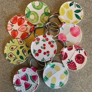 May include: A set of nine round keychains with different fruit designs. The keychains are white with colorful fruit illustrations, including strawberries, cherries, watermelon, pineapple, kiwi, avocado, raspberries, and lemons. Each keychain has a silver metal ring.