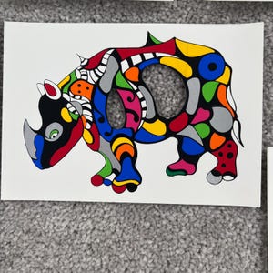 May include: A vibrant illustration of a rhinoceros. The rhino is rendered in a patchwork style, featuring a mix of colours such as red, blue, yellow, green, and grey. The artwork is set against a white backdrop.