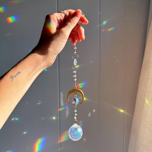May include: A gold-coloured moon and crystal suncatcher. The suncatcher features a crescent moon, iridescent crystals, and a large, clear crystal ball at the bottom. The suncatcher is suspended by a gold chain and ring.
