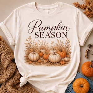 May include: A cream-coloured short-sleeve t-shirt featuring the brown text Pumpkin SEASON above a graphic illustration of various orange and white pumpkins nestled among dried autumn foliage and fallen leaves on the ground.