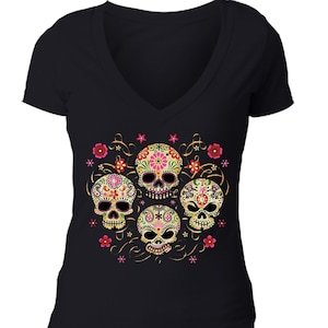 May include: Black V-neck t-shirt featuring a colorful Day of the Dead sugar skull design. The graphic includes five decorated skulls with floral and swirl accents in pink, yellow, and green.