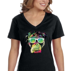 Könnte beinhalten: Ein schwarzes V-Ausschnitt-T-Shirt mit einem bunten Grafikdruck eines Schimpansen mit Sonnenbrille. Das Gesicht des Schimpansen ist mit Farbspritzern bemalt, darunter Grün, Blau, Gelb und Pink.