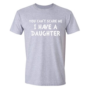 Puede incluir: Camiseta gris claro con el texto blanco "YOU CAN'T SCARE ME I HAVE A DAUGHTER". La camiseta es de cuello redondo y parece estar hecha de un material suave. Prenda casual.
