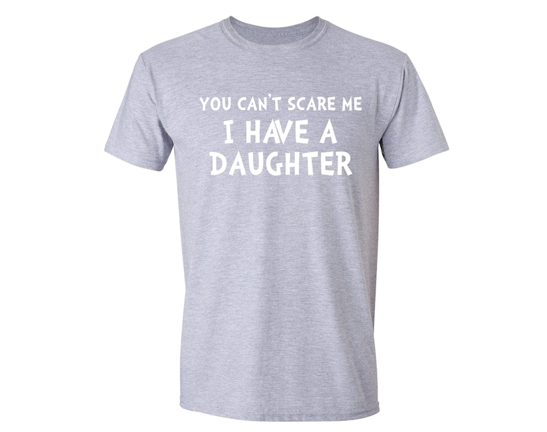 Puede incluir: Camiseta gris claro con el texto blanco "YOU CAN'T SCARE ME I HAVE A DAUGHTER". La camiseta es de cuello redondo y parece estar hecha de un material suave. Prenda casual.