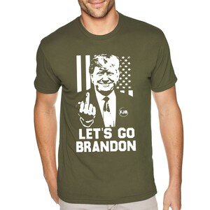 May include: A green t-shirt featuring a white image of a man giving the middle finger. The image is superimposed on an American flag. The text "Let's Go Brandon" is printed below the image.