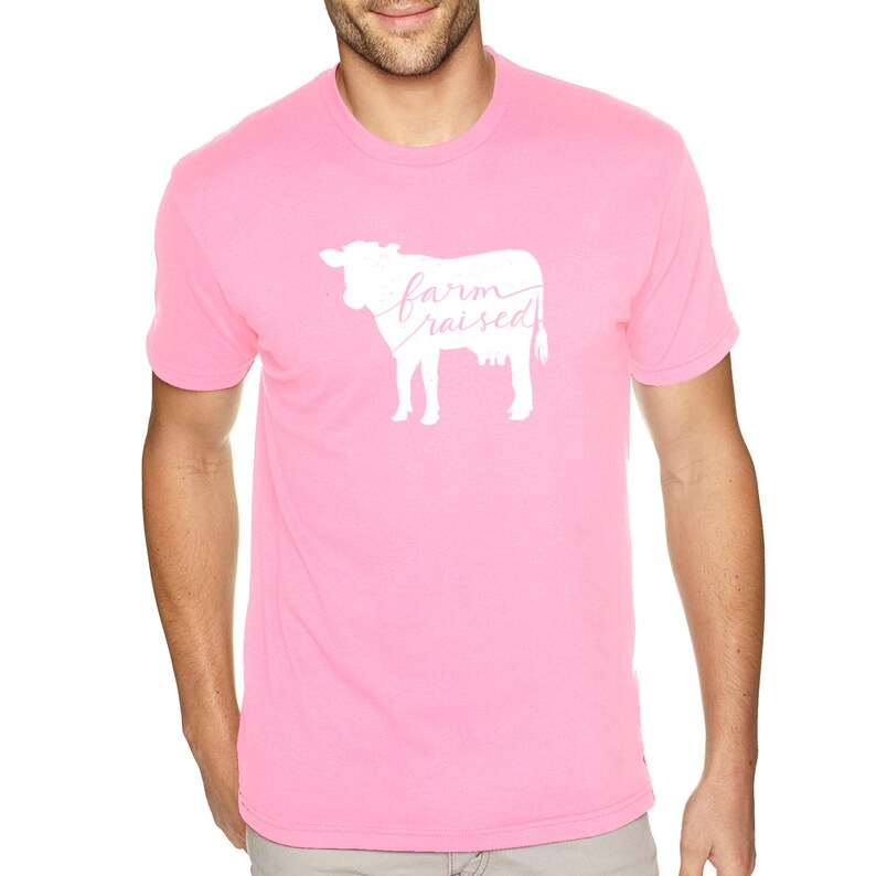 May include: Pink t-shirt featuring a white silhouette of a cow and the text "farm raised" in pink lettering.