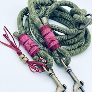 May include: An olive green rope dog leash featuring two antique brass swivel snap hooks, decorative dark red and pink thread whipping, and a matching red tassel accent with two small brass beads attached to the handle loop.