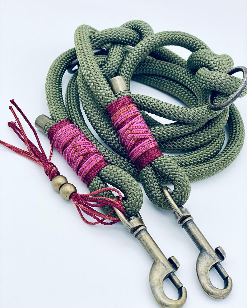 May include: An olive green rope dog leash featuring two antique brass swivel snap hooks, decorative dark red and pink thread whipping, and a matching red tassel accent with two small brass beads attached to the handle loop.