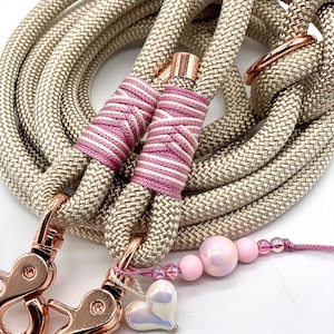 May include: A coiled beige rope leash featuring rose gold metal swivel clasps and hardware. The rope is secured with pink and white striped thread wrappings. A decorative charm strand with pink beads and a heart-shaped pendant is attached.