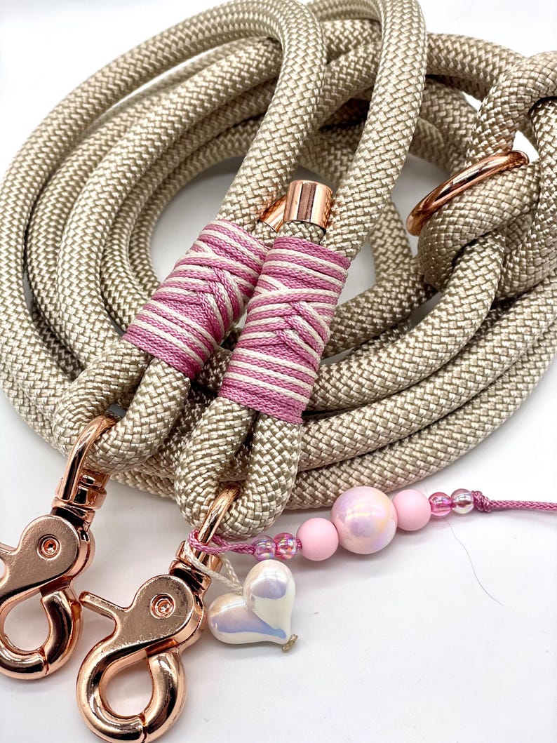 May include: A coiled beige rope leash featuring rose gold metal swivel clasps and hardware. The rope is secured with pink and white striped thread wrappings. A decorative charm strand with pink beads and a heart-shaped pendant is attached.