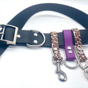 Puede incluir: Un cinturón negro con hebilla y anilla en D plateadas, con tres correas adjuntas. Dos correas tienen un diseño de estampado de leopardo y una es morada. Cada correa tiene un cierre o anillo de metal.