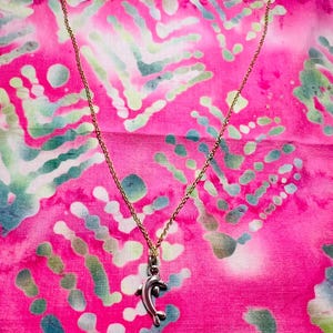 May include: A gold-colored chain necklace with a silver dolphin pendant. The dolphin pendant is detailed with fins and a curved body. The necklace is displayed on a vibrant pink and green patterned background.