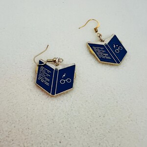 May include: A pair of gold-colored earrings featuring a blue book design. Each earring depicts an open book with white lines representing pages, and a lightning bolt and glasses symbol. The earrings have a hook closure.