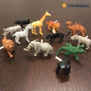 Könnte beinhalten: Ein Set mit 12 Tier-Schubladenknöpfen aus Kunststoff, mit einer Vielzahl von Tieren, darunter ein Löwe, ein Zebra, eine Giraffe, ein Elefant, ein Nashorn, ein Kamel, ein Gorilla, ein Krokodil, ein Flusspferd, ein Panther, ein Tiger und ein Bär. Die Knöpfe sind alle in verschiedenen Farben gehalten und haben ein realistisches Design.