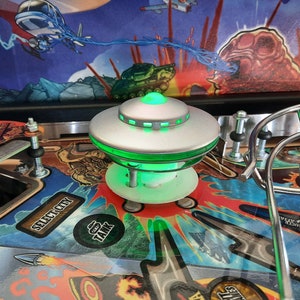May include: A green and silver UFO pinball machine toy with a glowing green light. The machine has a black and red background with a cartoon image of a monster.