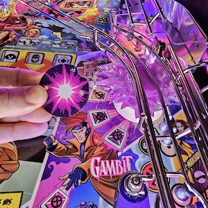 May include: A close-up of a pinball machine with a purple and pink background. The machine features a character named Gambit, who is throwing a playing card. The card is a purple and pink diamond with a symbol in the center. The machine also features a score of -13.