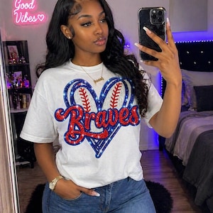 Puede incluir: Camiseta blanca con un diseño de béisbol en forma de corazón y la palabra "Braves" en purpurina roja y azul. La camiseta se combina con vaqueros azules. Un letrero de neón que dice "Good Vibes" es visible en el fondo.