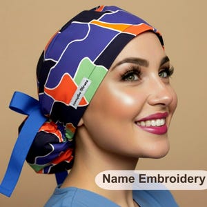 May include: A scrub cap featuring a geometric pattern of blue, orange, black, and light green shapes. The cap has a blue ribbon tie at the back and a small white tag with the text Everyday Heroes. The text Name Embroidery appears in the corner.