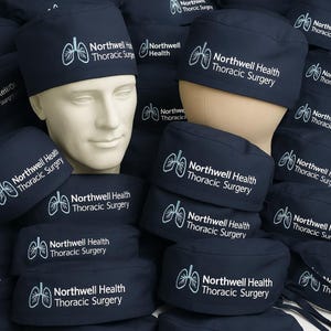 Puede incluir: Una colección de gorros quirúrgicos azul marino con un gráfico bordado en blanco de pulmones humanos y el texto Northwell Health Thoracic Surgery. Los gorros se muestran sobre cabezas de maniquí y apilados para mayor visibilidad.