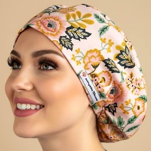 May include: A scrub cap featuring a light pink background with a floral pattern of pink, yellow, and dark green leaves and flowers. The cap has a white fabric label on the side with the text "scrubby heroes" printed in a dark font.