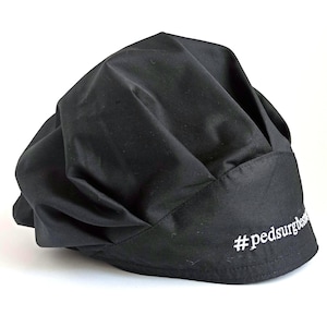 May include: A solid black scrub cap featuring a gathered, voluminous crown and a structured front band. The text #pedsurgbest is embroidered in white thread on the right side of the band. The fabric has a matte finish with visible folds and pleats.