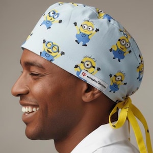 Op de afbeelding: Een lichtblauwe chirurgische scrubcap met een herhaald patroon van gele Minion-personages in verschillende poses. De cap heeft een gele stropdas en de tekst "everyday Hanna" op een wit label.