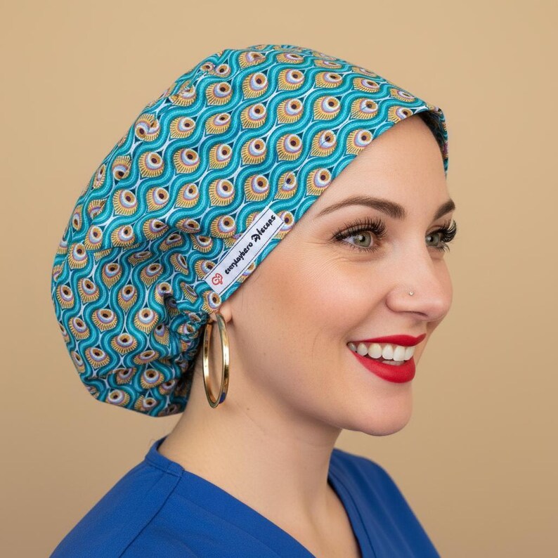 Woman in 3/4 angle wearing turquoise peacock feather scrub cap with green hazel eyes and gold hoop earrings - vibrant teal bouffant surgical hat with peacock eye pattern in gold, orange and purple - euro style medical headwear with everydayheroescaps label - professional scrub cap for nurses and healthcare workers