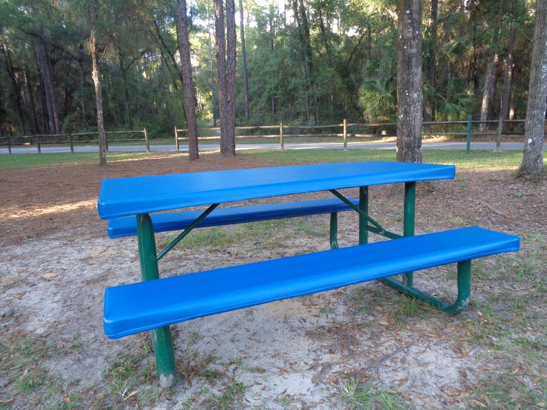 May include: A blue picnic table with a metal frame and a blue cover. The table is in a grassy area with trees in the background.