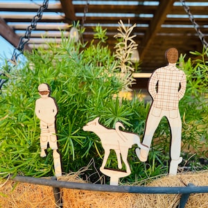 May include: Three wooden figures, a person, a dog, and another person, are placed in a hanging planter filled with green plants. The figures are painted in shades of brown and white.