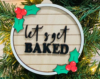 May include: A round wooden Christmas ornament with a white background and a wood grain pattern. It has green holly leaves and red berries on either side of the text "Let's get baked".