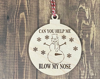 May include: A wooden Christmas ornament shaped like a round ball with a snowman design. The ornament has the text "Can you help me blow my nose" and a snowman with a carrot nose.