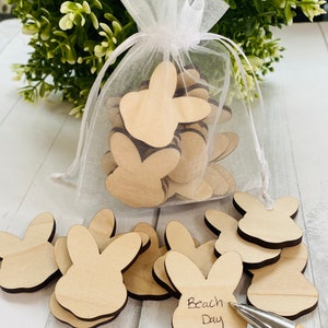 May include: A white organza bag filled with wooden bunny-shaped tags. Some tags are scattered on a white wooden surface, one of which has the words "Beach Day" written on it.
