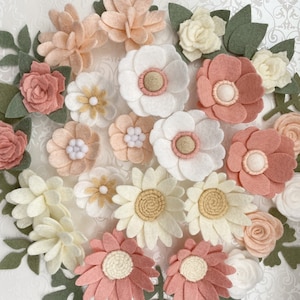 May include: A collection of felt flower embellishments in various shades of pink, white, and cream. The flowers are arranged in a circular pattern and include different sizes and styles, such as daisies, roses, and simple round flowers.