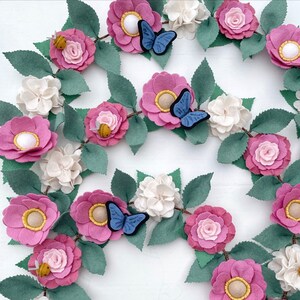 May include: A felt garland with pink, white, and green flowers, butterflies, and bees. The garland is perfect for decorating a nursery, bedroom, or party.