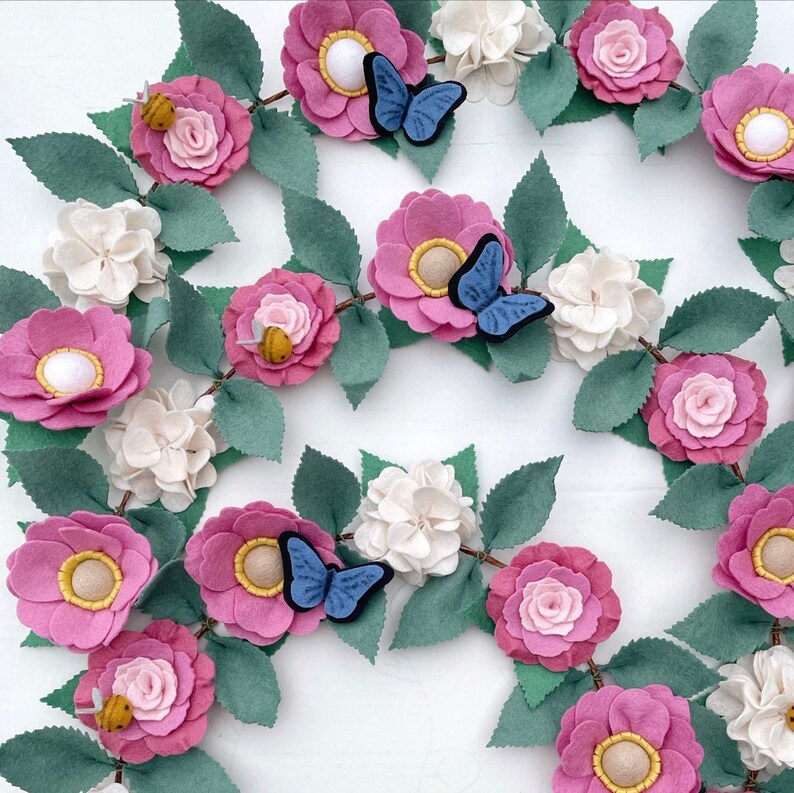 May include: A felt garland with pink, white, and green flowers, butterflies, and bees. The garland is perfect for decorating a nursery, bedroom, or party.