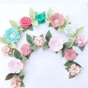 May include: A garland of felt flowers in shades of pink, light green, and turquoise. The garland is made up of large flowers and smaller hydrangea-like flowers. The flowers are strung together with green felt leaves.