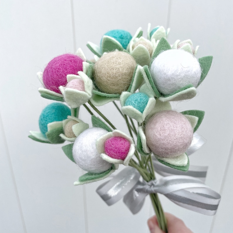 May include: A bouquet of felt flowers with a variety of colors, including pink, white, beige, and turquoise. The flowers are tied together with a silver ribbon.