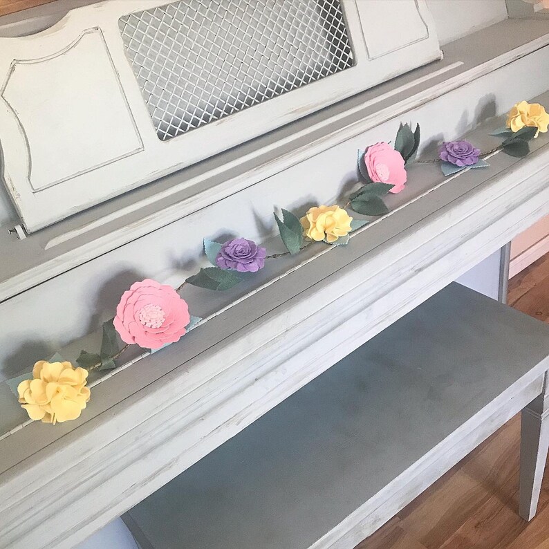May include: A garland of felt flowers in pink, yellow, and purple, with green leaves, is draped across the top of a light gray piano.