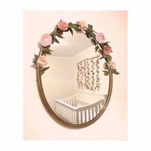 May include: A gold oval mirror with a floral garland made of pink felt flowers and green leaves. The mirror is reflecting a white crib and a wall with a floral garland.