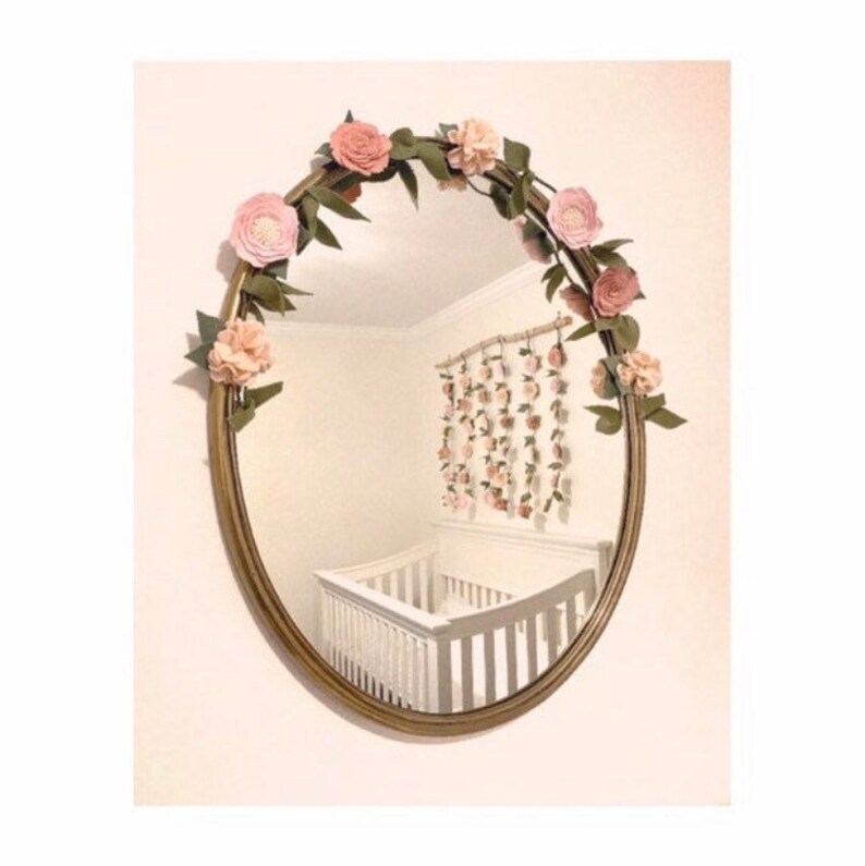 May include: A gold oval mirror with a floral garland made of pink felt flowers and green leaves. The mirror is reflecting a white crib and a wall with a floral garland.