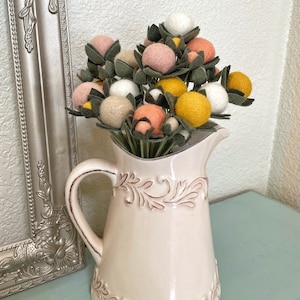 May include: A bouquet of felt flowers in a white ceramic pitcher. The flowers are pink, yellow, and white and have green leaves.