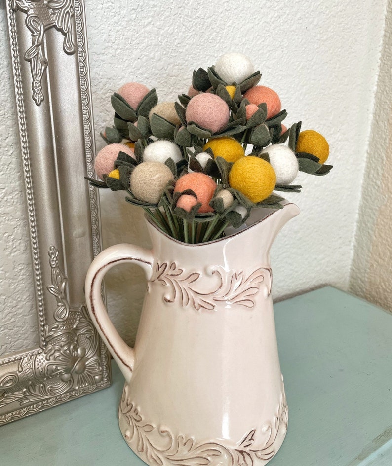 May include: A bouquet of felt flowers in a white ceramic pitcher. The flowers are pink, yellow, and white and have green leaves.
