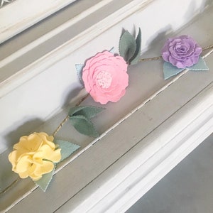May include: A felt flower garland with pink, yellow, and purple flowers and green leaves. The garland is strung on twine and is perfect for decorating a nursery or children's room.
