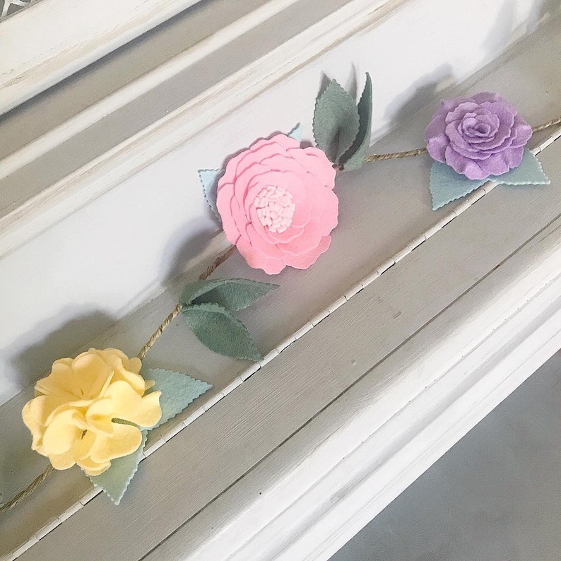May include: A felt flower garland with pink, yellow, and purple flowers and green leaves. The garland is strung on twine and is perfect for decorating a nursery or children's room.