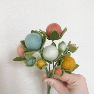 May include: A bouquet of felt flowers with green leaves and pastel colored felt balls. The flowers are arranged in a hand.
