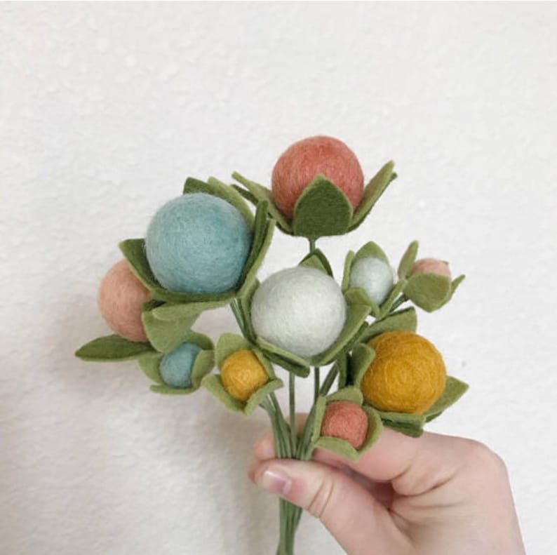 May include: A bouquet of felt flowers with green leaves and pastel colored felt balls. The flowers are arranged in a hand.