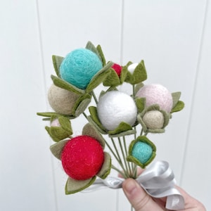 May include: A bouquet of felt flowers with green leaves and colorful felt balls in shades of pink, white, red, and blue. The bouquet is tied with a white ribbon.